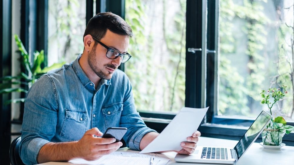 Serious pensive thoughtful focused young casual entrepreneur small business owner accountant bookkeeper in office looking at and working with laptop and income tax return papers and documents