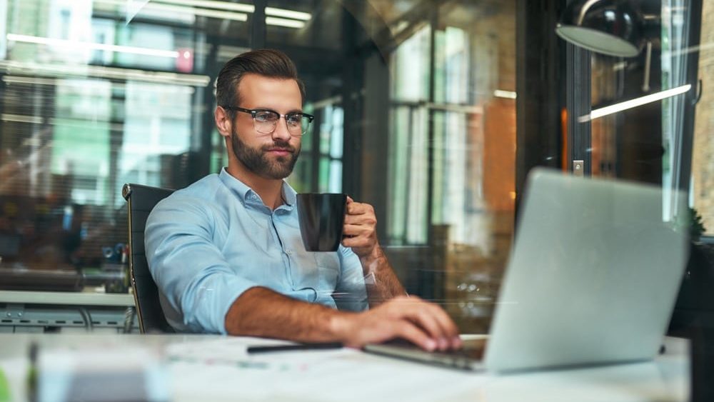 Morning coffee. Portrait of young and successful bearded man in eyeglasses holding cup of coffee and working with laptop while sitting at his working place